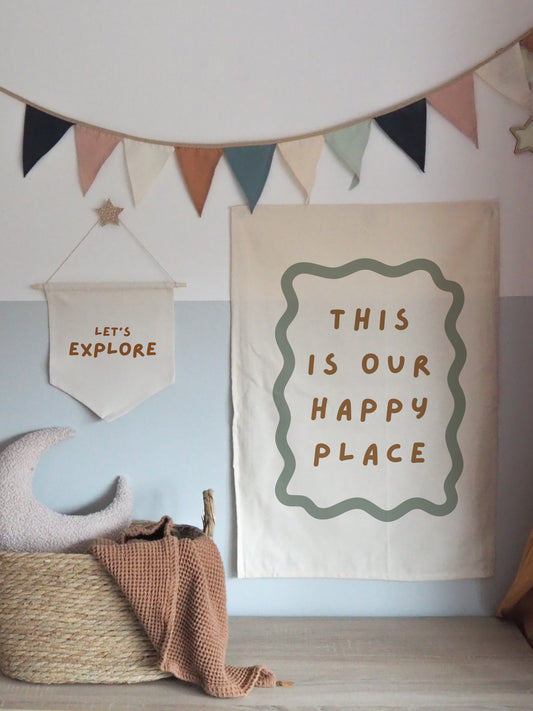A banner with the text 'THIS IS OUR HAPPY PLACE' displayed at a children's room decor setting, featuring a beige background with green lettering, accompanied by children's toys and decorative flags.